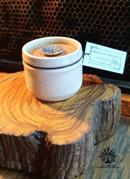 Pot de baume artisanal à la cire d'abeille posé sur une souche de bois, avec en arrière-plan un motif en nid d'abeille, étiqueté "BAUME DU FORGERON À l'Huile d'Olive, à la Cire d'Abeille"