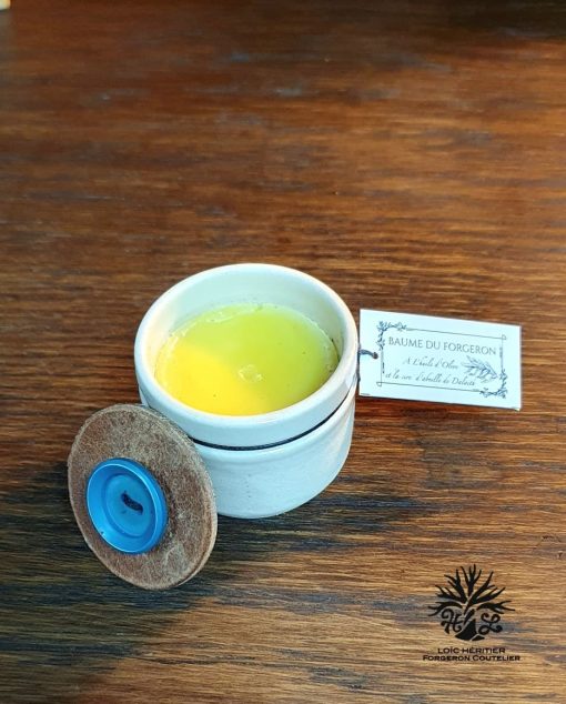 Un pot de baume artisanal ouvert, avec un étiquetage manuscrit sur une table en bois, à côté d'une étiquette indiquant "BAUME DU FORGERON À l'Huile d'Olive, à la Cire d'Abeille", fabriqué par Loïc Héritier, coutelier.